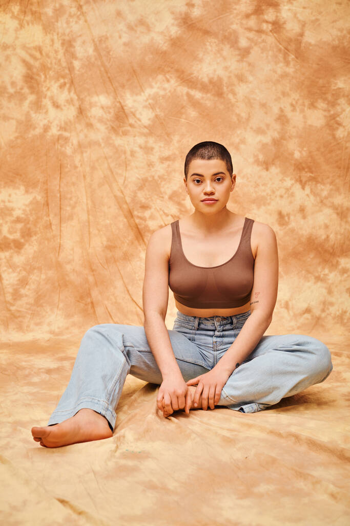 body positivity, denim fashion, curvy tattooed woman in jeans and crop top sitting on mottled beige background, casual attire, looking at camera, self-acceptance, generation z, short haired model  - Photo, Image