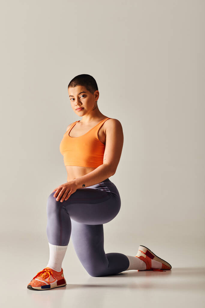 движение тела позитивности, молодая коротковолосая женщина стоя на коленях на сером фоне, кривая модель фитнеса в спортивной одежде, расширение прав и возможностей, мотивация, тренировка с поднятыми руками, сила и здоровье  - Фото, изображение