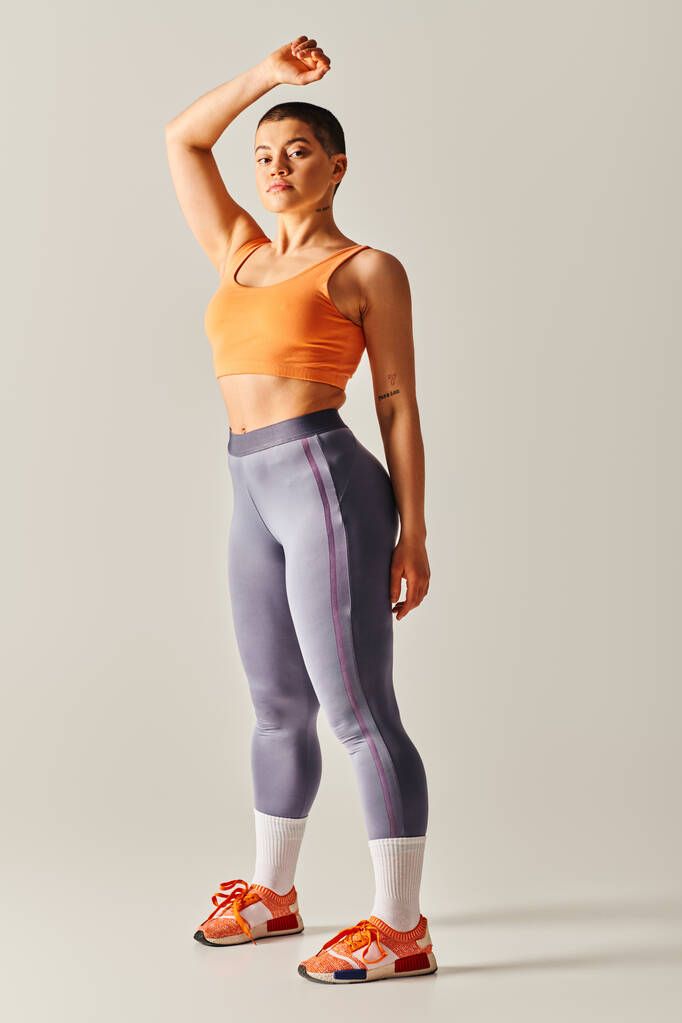 sebevědomí těla, atletická a krátkosrstá žena pózující na šedém pozadí, zakřivený fitness model, stojící se zvednutou rukou, vytrvalost a posílení postavení, generace z, plná délka  - Fotografie, Obrázek