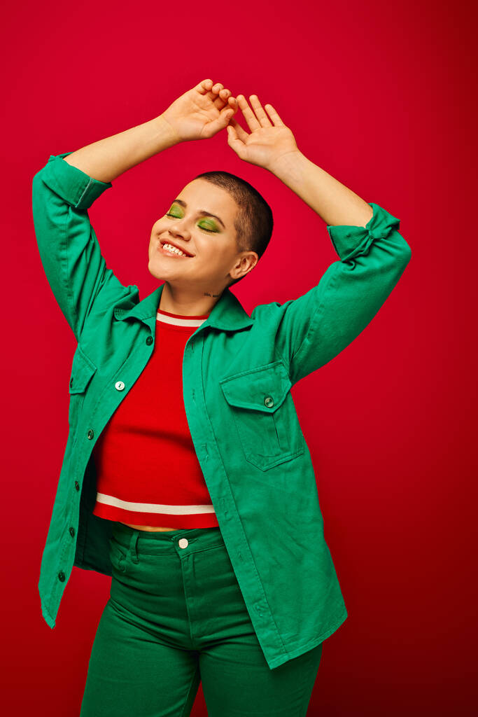 fashion and style, happy and short haired woman in green outfit posing with raised hands on red background, smiling, generation z, youth culture, modern backdrop, individuality, personal style  - Photo, Image