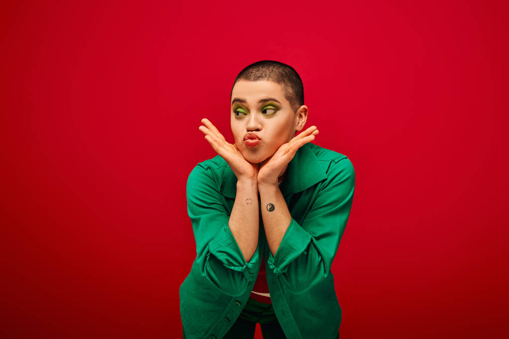 móda a styl, emocionální a tetované, krátkosrstá žena v zeleném oblečení trucuje rty na červeném pozadí, dívá se jinam, generace z, mládež kultura, pulzující pozadí, půvab  - Fotografie, Obrázek