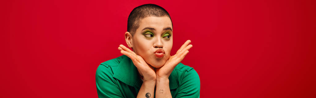 moda e estilo, emocional e tatuado, mulher de cabelos curtos em roupa verde bebendo lábios em fundo vermelho, olhando para longe, geração z, cultura da juventude, pano de fundo vibrante, individualidade, banner - Foto, Imagem