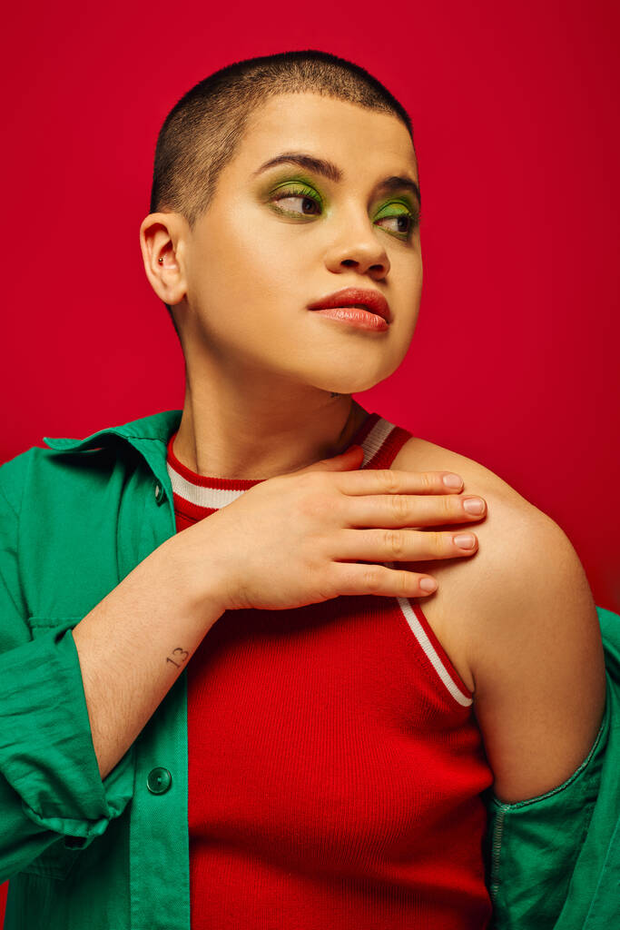 muotisuuntaus, nuorekas ja tatuoitu, lyhytkarvainen nainen vihreässä asussa poseeraa punaisella taustalla, katselee pois, sukupolvi z, nuoriso, elinvoimainen tausta, rohkea meikki, henkilökohtainen tyyli, muotokuva  - Valokuva, kuva