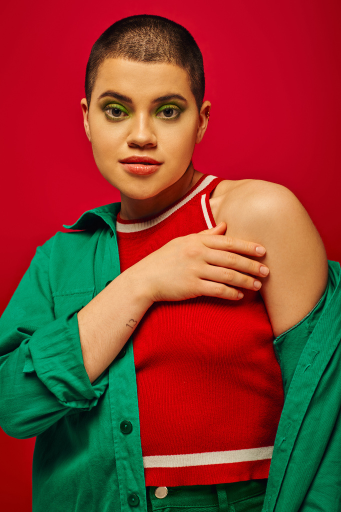 fashion trend, youthful and tattooed, short haired woman in green outfit posing on red background, looking at camera, generation z, youth, vibrant backdrop, bold makeup, personal style, portrait  - Photo, Image