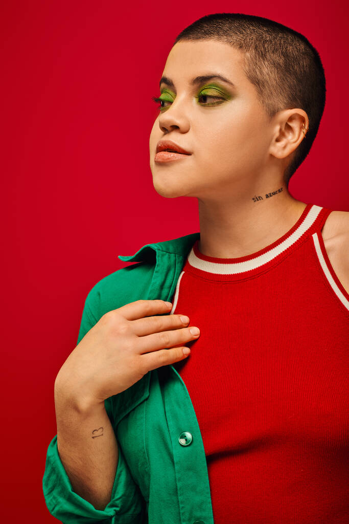 Modetrend, jung und tätowiert, kurzhaarige Frau im grünen Outfit posiert auf rotem Hintergrund, schaut weg, Generation Z, Jugend, lebendige Kulisse, fettes Make-up, Porträt  - Foto, Bild