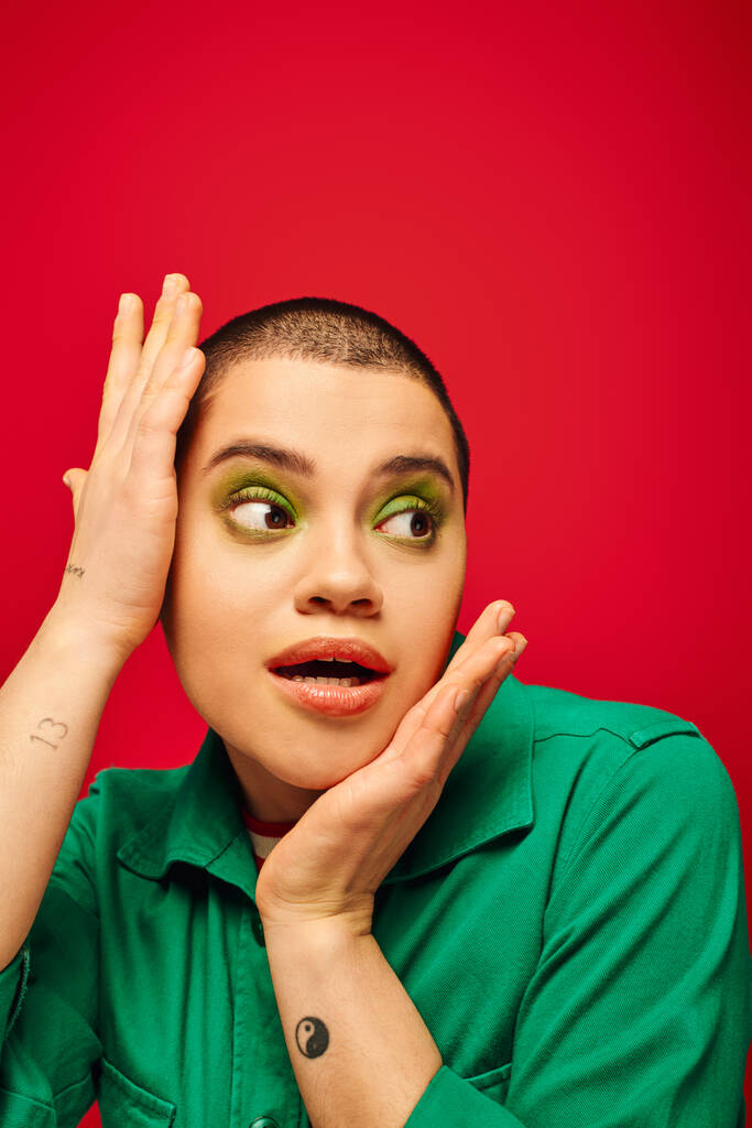 fashion choices, amazed and tattooed, short haired woman in green outfit touching head on red background, looking away, generation z, youth, vibrant backdrop, individuality, personal style  - Photo, Image