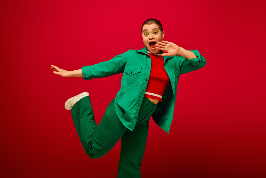 stylish look, amazed and tattooed, short haired woman in green outfit gesturing on red background, looking at camera, generation z, youth, vibrant backdrop, individuality, personal style  - Photo, Image