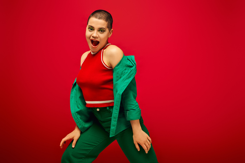 stylish appearance, bold makeup, amazed and tattooed, short haired woman in green outfit posing on red background, generation z, youth, vibrant backdrop, individuality, personal style  - Photo, Image