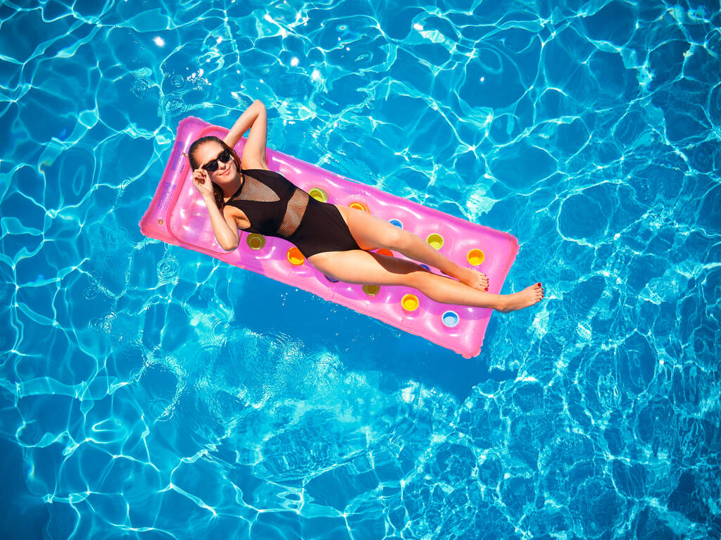 Fit pretty girl in bikini chilling on inflatable pink mattress in swimming pool. Slim hot woman in swimwear tanning. Female relaxing on a float in blue water at luxury resort. Aerial, view from above. - Photo, Image