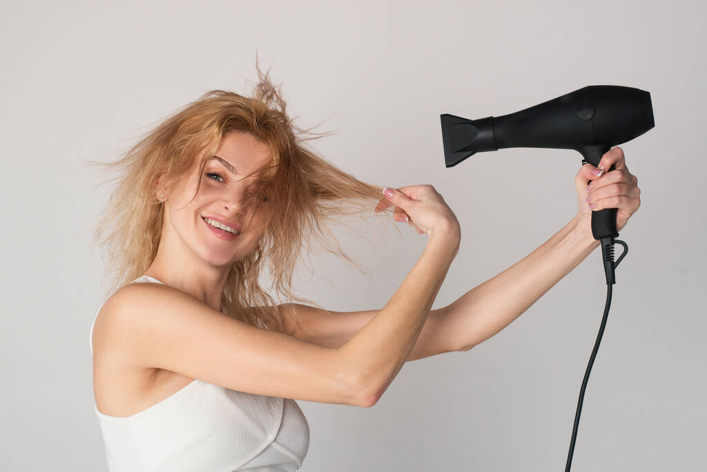 Krásná běloška pózuje s mokrými vlasy. Léčba ženských vlasů. Dívka s blond vlasy pomocí fénu. účes, koncept kadeřnictví - Fotografie, Obrázek