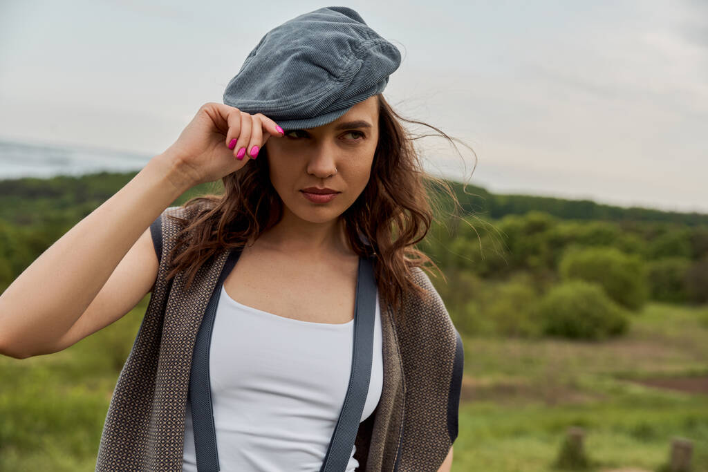 Fashionable brunette woman in suspenders and vintage clothes wearing newsboy cap and looking away while standing with blurred nature at background, fashion-forward in countryside - Photo, Image