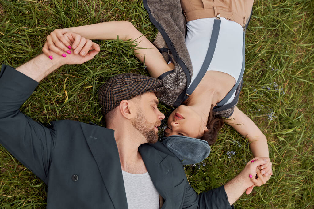 Top view of trendy and bearded man in newsboy cap and vintage jacket holding hands of stylish girlfriend in susenders while lying on grassy lawn, fashion-forward in countryside - Фото, изображение