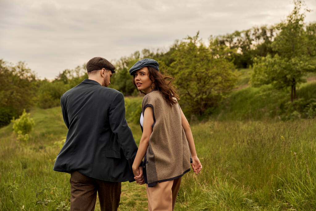Fashionable brunette woman in vest and newsboy cap looking at camera and holding hand of bearded boyfriend and walking with landscape at background, stylish couple in rural setting - Photo, Image