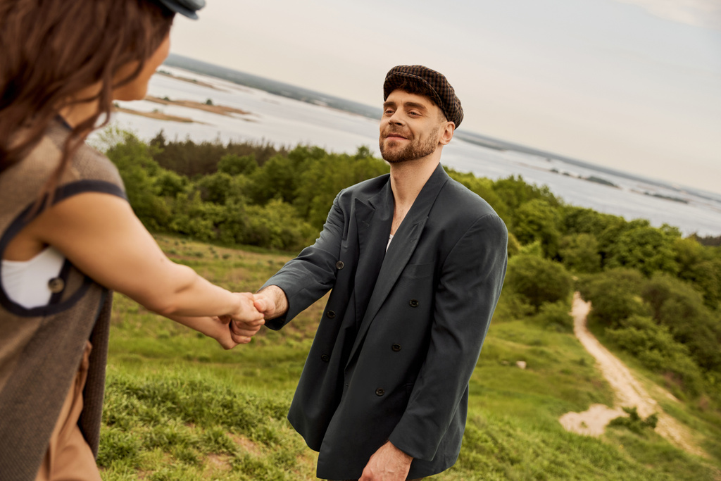 Smiling and bearded man in stylish newsboy cap and jacket holding hand of blurred brunette girlfriend while standing with blurred nature and sty at background, fashion-forwards in countryside - Photo, Image