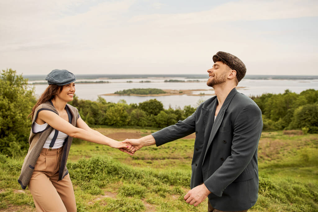 Pozitivní a módní brunetka žena v newsboy čepice a vesta drží ruku vousatého přítele v bundě, zatímco stojí s malebnou krajinou na pozadí, módní pár na venkově - Fotografie, Obrázek