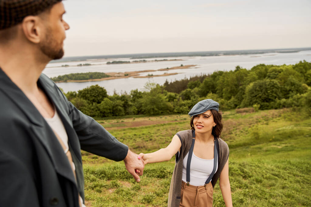 Smiling and trendy brunette woman in suspenders and newsboy cap holding hand of blurred boyfriend in jacket while standing with nature and sky at background, fashionable couple in countryside - Photo, Image