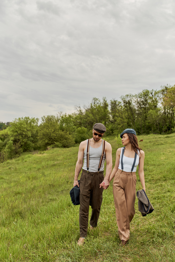 Fashionable brunette woman in newsboy cap and suspenders holding hand of bearded boyfriend in sunglasses and talking while walking together on grassy lawn, trendy couple in the rustic outdoors - Photo, Image