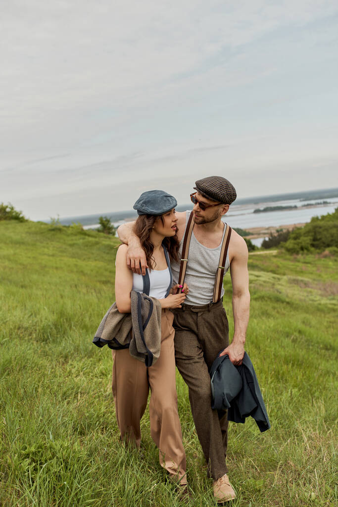 Modny mężczyzna w zabytkowym stroju i okularach przeciwsłonecznych przytulając brunetkę i rozmawiając podczas spaceru po rozmazanym trawiastym wzgórzu z zachmurzonym niebem na tle, stylowi partnerzy w wiejskiej ucieczce - Zdjęcie, obraz