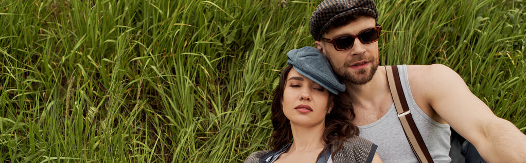 Stylish brunette woman in newsboy cap sitting with closed eyes next to bearded boyfriend in sunglasses and vintage outfit on grassy hills, fashionable couple surrounded by nature, banner  - Photo, Image