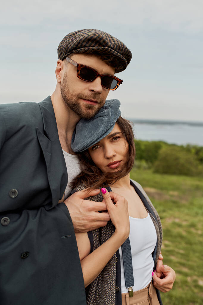 Πορτρέτο της μόδας άνθρωπος σε γυαλιά ηλίου και σακάκι αγκαλιάζει μελαχρινή φίλη σε newsboy καπέλο και vintage ρούχα και κοιτάζοντας κάμερα με θολή τοπίο στο παρασκήνιο, ρομαντική απόδραση - Φωτογραφία, εικόνα