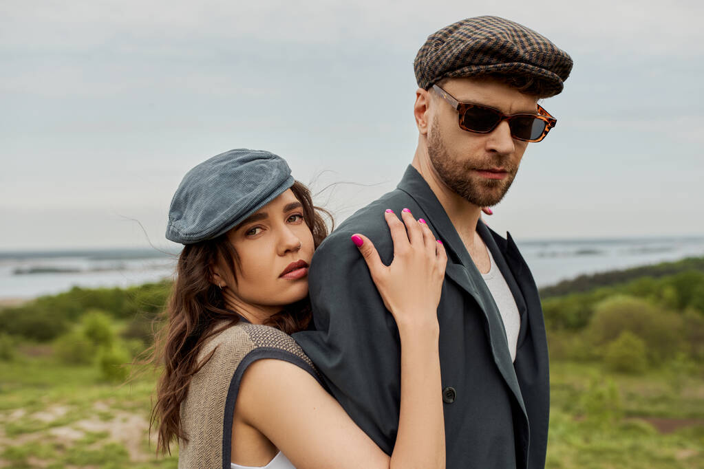 Portrait of trendy brunette woman in newsboy cap and vest hugging boyfriend in sunglasses and jacket while looking at camera with landscape at background, trendy twosome in rustic setting - Photo, Image