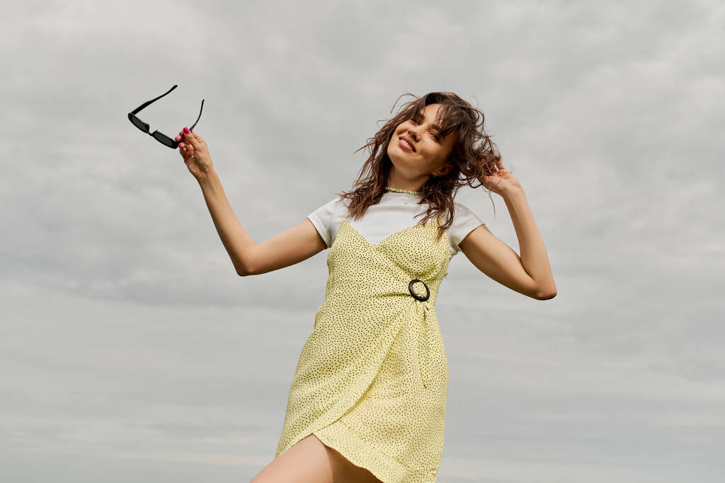 Low angle view of cheerful and stylish brunette woman in sundress looking at camera and holding sunglasses while standing with cloudy sky at background, summertime joy - Photo, Image