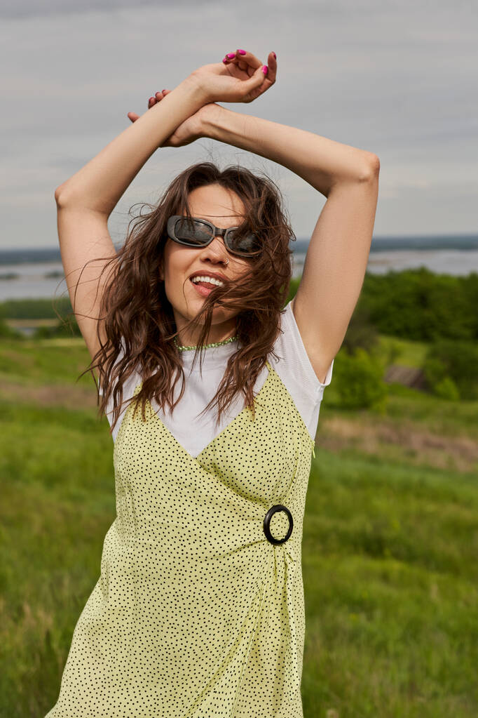 Retrato de mujer morena de moda y alegre en gafas de sol y vestido de sol posando mientras está de pie con el paisaje natural borroso y el cielo al fondo, alegría de verano - Foto, Imagen