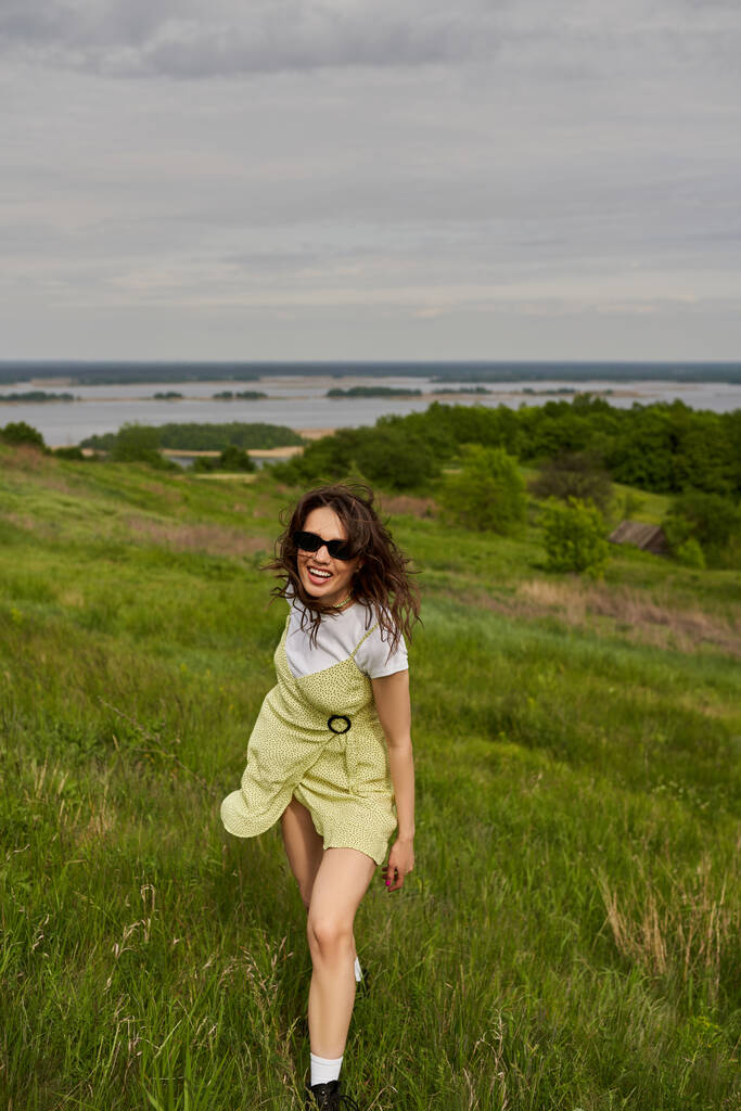 Femme brune joyeuse et élégante en lunettes de soleil et robe de soleil debout sur la prairie verte avec de l'herbe et passer du temps avec un paysage flou et le ciel en arrière-plan, joie estivale - Photo, image