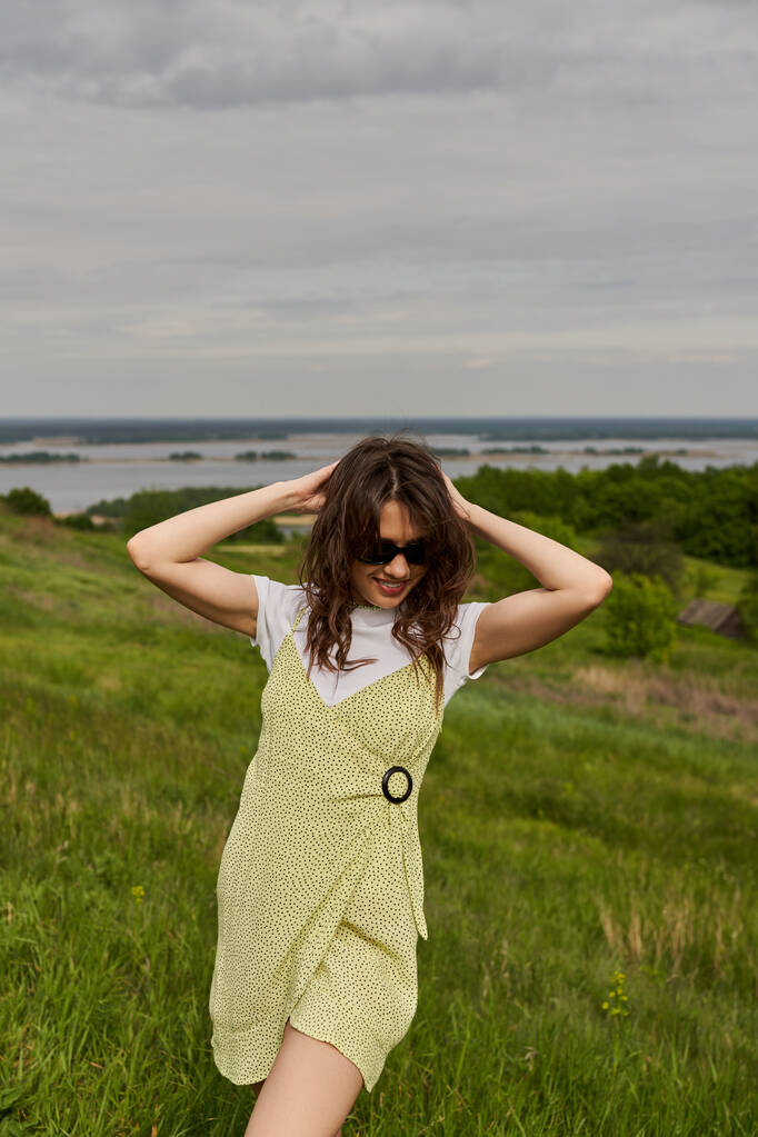 Trendy and smiling brunette woman in sunglasses and stylish sundress touching head and standing while spending time on blurred grassy meadow at background, summertime joy - Photo, Image