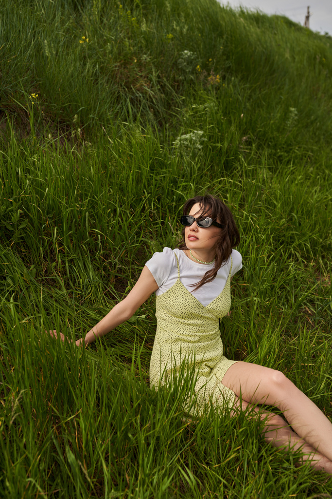 Mulher morena elegante em óculos de sol e vestido de sol relaxante e passar o tempo sentado na colina com grama verde no fundo, paisagem natural e conceito de espírito livre - Foto, Imagem
