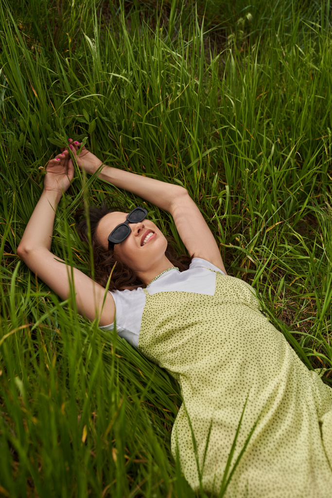 Vista superior de mujer morena alegre y elegante en gafas de sol y vestido de sol tumbado y relajante en el césped cubierto de hierba en verano, paisaje natural y relajante en concepto de naturaleza, paisaje rural - Foto, imagen