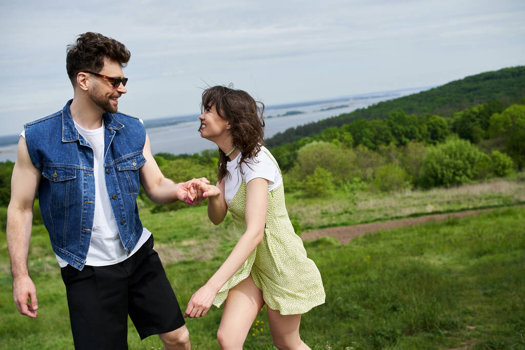 Smiling and stylish man in sunglasses holding hand of cheerful girlfriend in sundress while spending time and standing together with scenic landscape at background, couple in love enjoying nature - Photo, Image