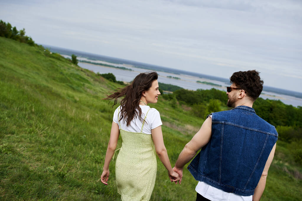 Seitenansicht des lächelnden und trendigen romantischen Paares im Sommer-Outfit Händchen haltend auf grasbewachsenen Hügeln mit bewölktem Himmel im Hintergrund, verliebtes Paar, das die Natur genießt, Ruhe - Foto, Bild
