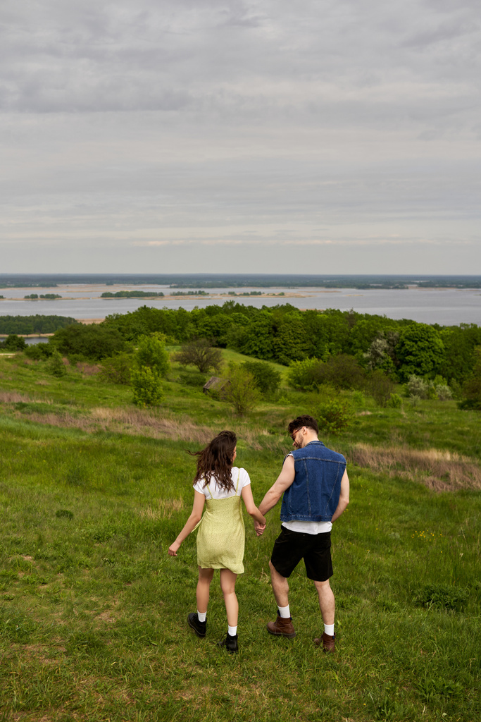 Plná délka stylové brunetky romantický pár v letních oblečení drží za ruce při relaxaci a chůzi na zeleném kopci s malebnou krajinou na pozadí, dobrodružství na venkově a milostný příběh - Fotografie, Obrázek
