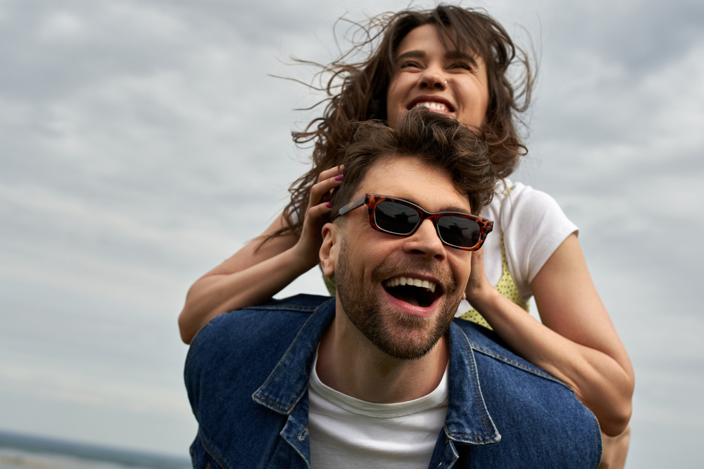 Cheerful brunette woman piggybacking on stylish boyfriend in sunglasses and denim vest and having fun together with cloudy sky at background, countryside adventure and love story - Photo, Image