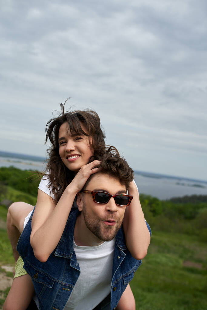 Cheerful brunette woman touching hair of excited and stylish boyfriend in sunglasses while piggybacking and having fun with scenic nature at background, countryside adventure and love story - Photo, Image