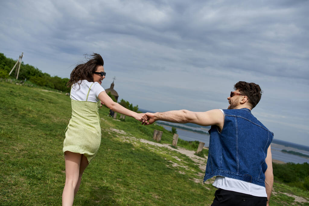 Pohled na pozitivní romantický pár v slunečních brýlích a letních šatech držící se za ruce při běhu na travnatém kopci s rozmazanou krajinou na pozadí, venkovskou touhou a konceptem lásky - Fotografie, Obrázek