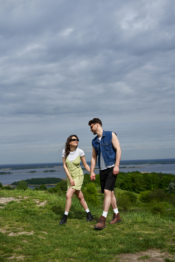 Positive brunette romantic couple in sunglasses and stylish summer outfits holding hands and walking together on grassy hill with blurred scenic view at background, countryside leisurely stroll - Photo, Image