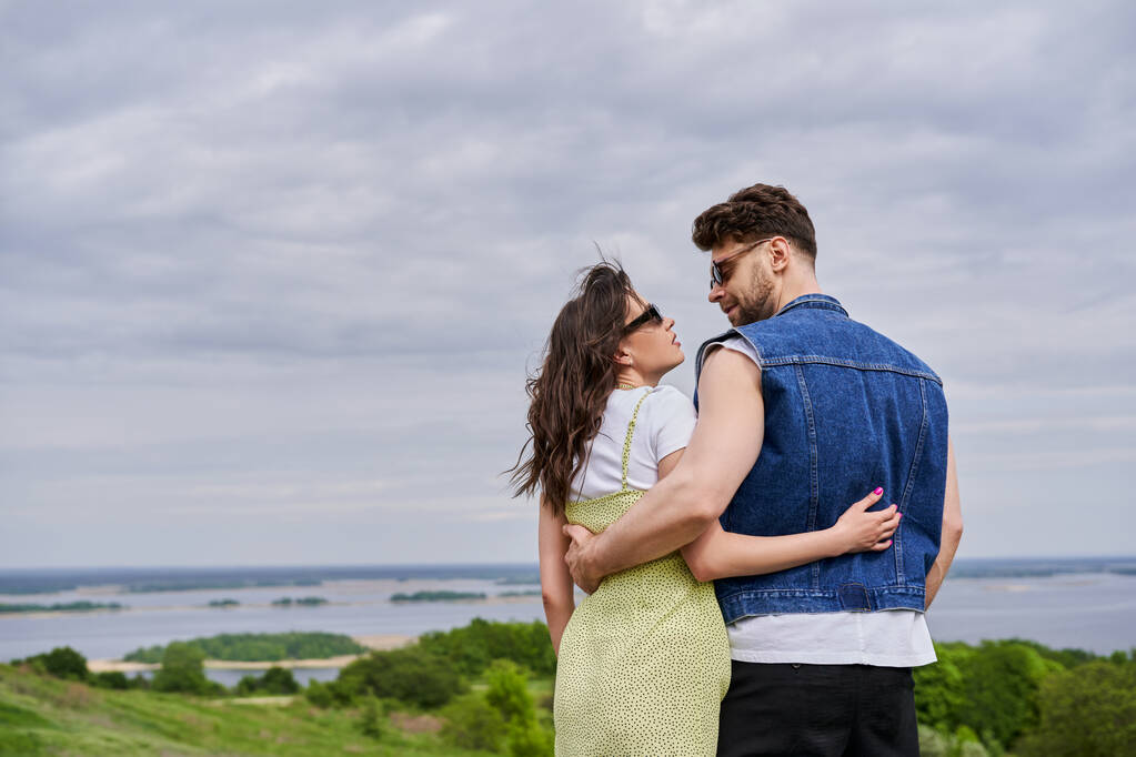 Side view of trendy romantic couple in sunglasses and summer outfits hugging and looking at each other with scenic landscape and cloudy sky at background, countryside leisurely stroll - Photo, Image