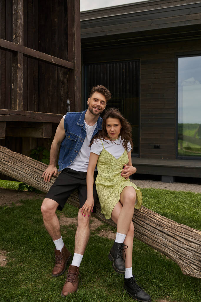 Veselý a stylový brunetka romantický pár v létě oblečení a boty objímání při sezení na dřevěné klády ve venkovském prostředí na pozadí, krajina ústup koncept, klid - Fotografie, Obrázek