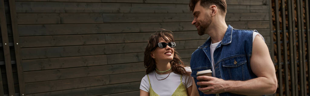 Uśmiechnięta brunetka w okularach przeciwsłonecznych i letnim stroju patrząc na brodatego chłopaka trzymającego kawę na wynos podczas spaceru w pobliżu drewnianego domu w tle, koncepcja przyjemności na świeżym powietrzu, baner  - Zdjęcie, obraz
