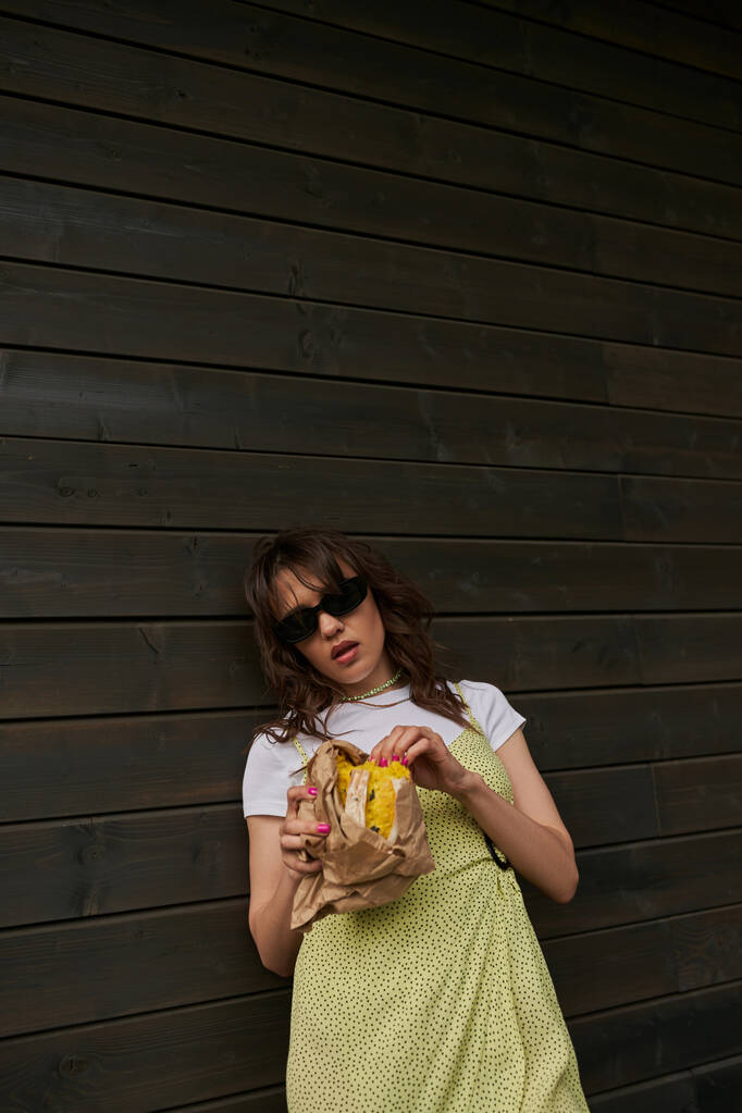Μοντέρνο μελαχρινή γυναίκα σε γυαλιά ηλίου και καλοκαιρινό ντύσιμο κρατώντας φρέσκο κουλούρι σε συσκευασία χειροτεχνίας, ενώ στέκεται κοντά στο ξύλινο σπίτι στο παρασκήνιο, το καλοκαίρι vibes έννοια, ηρεμία - Φωτογραφία, εικόνα