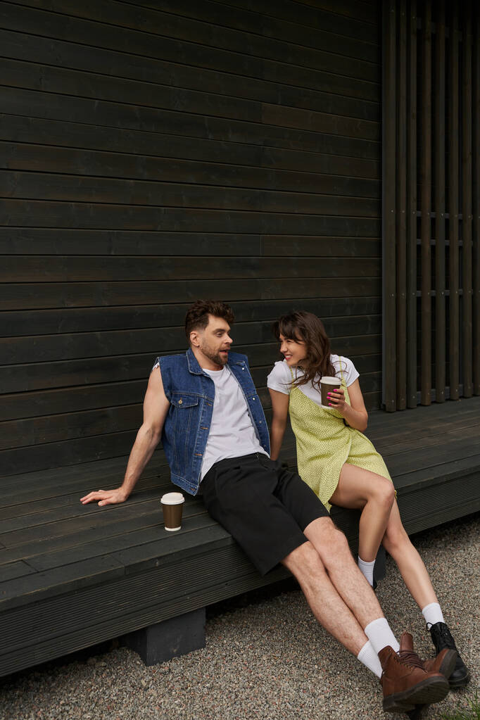 Pleine longueur de femme brune gaie dans une tenue d'été élégante tenant du café pour aller parler à son petit ami tout en étant assis ensemble près d'une maison en bois dans un cadre rural, concept de moments insouciants - Photo, image