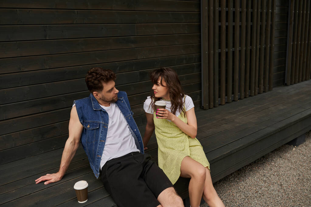 Stilvolle brünette Frau in Sonnenanzüge mit Kaffee zum Mitnehmen und Blick auf bärtigen Freund in Jeansweste, während sie in der Nähe von Holzhaus in ländlicher Umgebung sitzt, unbeschwerte Momente Konzept - Foto, Bild