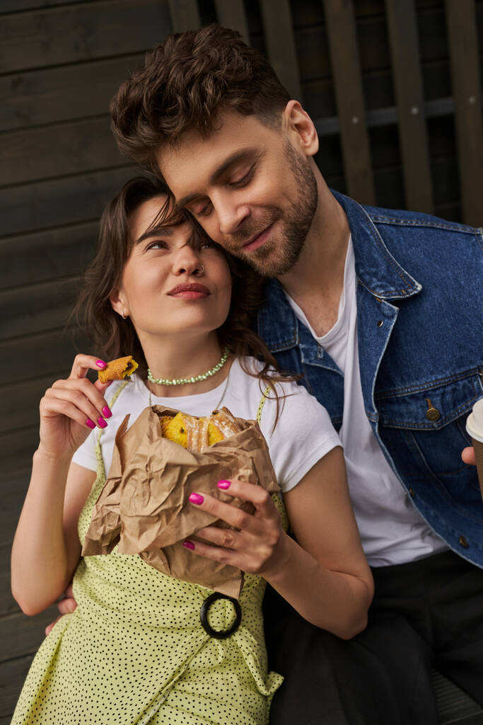 Portrait of joyful brunette woman in sundress holding fresh bun in craft package and looking at stylish boyfriend in denim vest while sitting near wooden house at background, serene ambiance concept - Photo, Image
