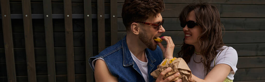 Весела брюнетка в сонцезахисних окулярах годує стильного хлопця зі свіжою булочкою, проводячи час біля дерев'яного будинку на задньому плані в сільській місцевості, безтурботну концепцію атмосфери, банер  - Фото, зображення