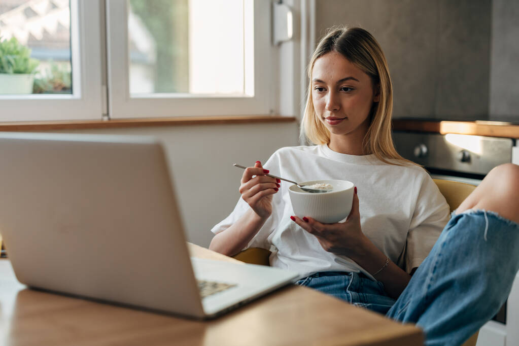 Jonge vrouw eet havermout in de keuken en kijkt naar de laptop. - Foto, afbeelding