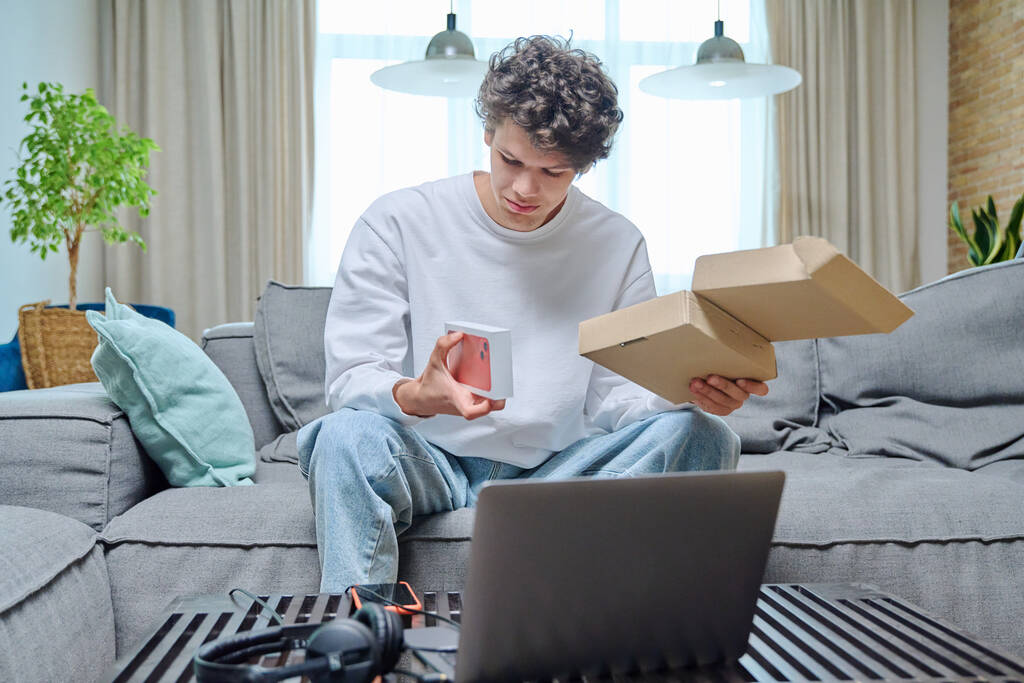 Genç bir erkek online alışveriş paketini açıyor, evdeki koltukta yeni akıllı telefonlu bir kutuyla oturuyor. İnternet alışverişi, dağıtım, gençlik kavramı - Fotoğraf, Görsel
