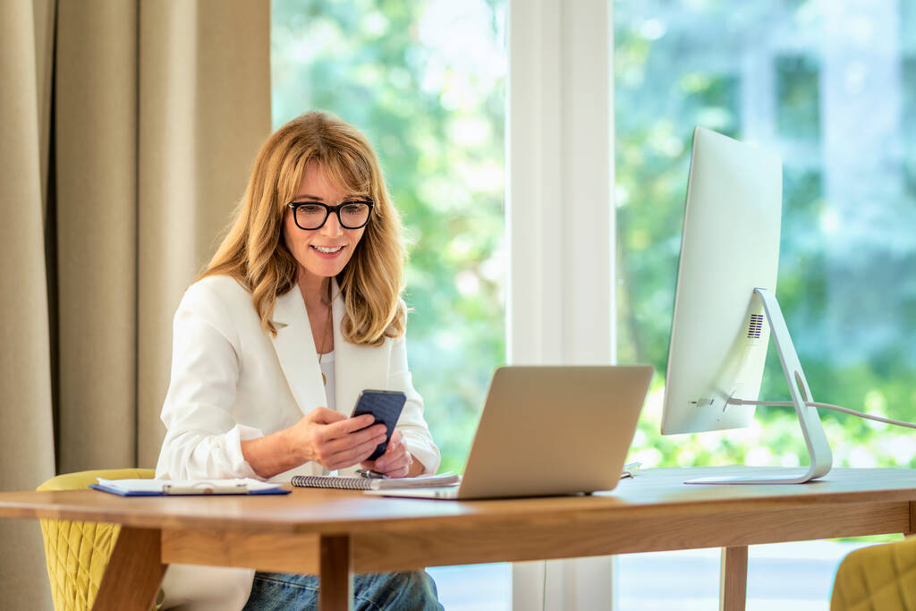 Fotografía de una mujer de mediana edad sentada en el escritorio y usando el teléfono móvil. Mujer segura con gafas y chaqueta blanca mientras trabaja desde casa. Oficina en casa. - Foto, Imagen