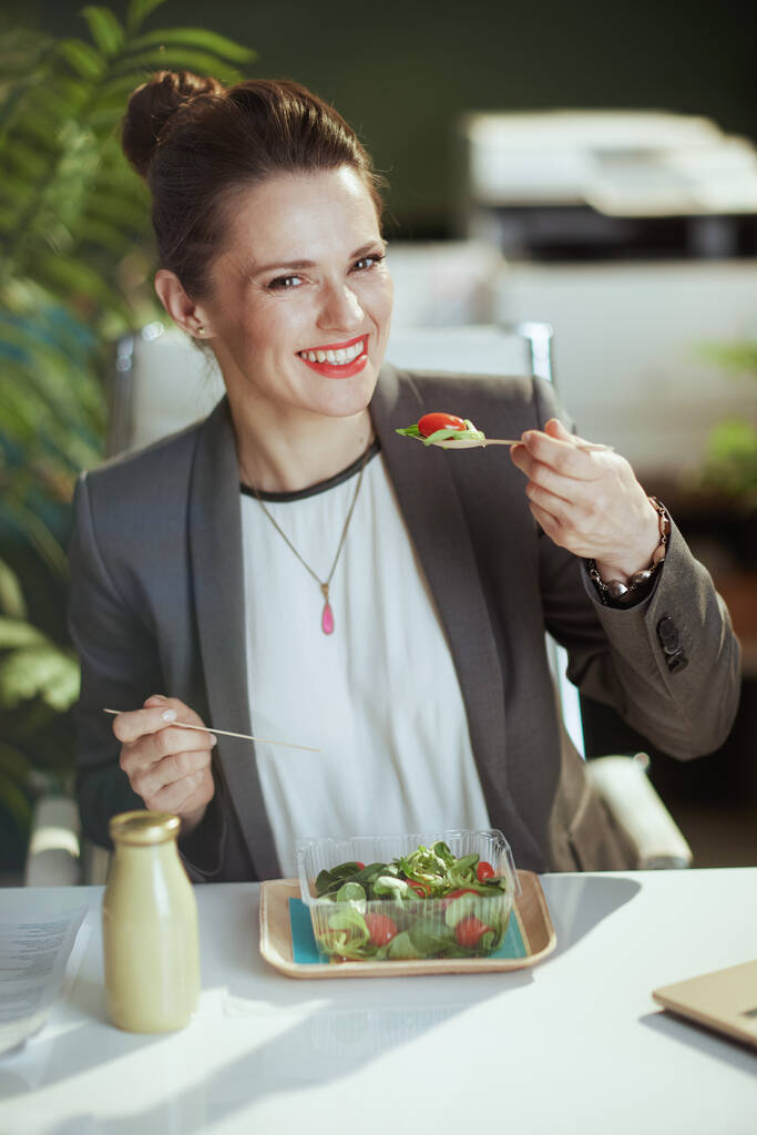 Posto di lavoro sostenibile. felice moderno lavoratore donna di mezza età in un abito grigio business in moderno ufficio verde mangiare insalata. - Foto, immagini